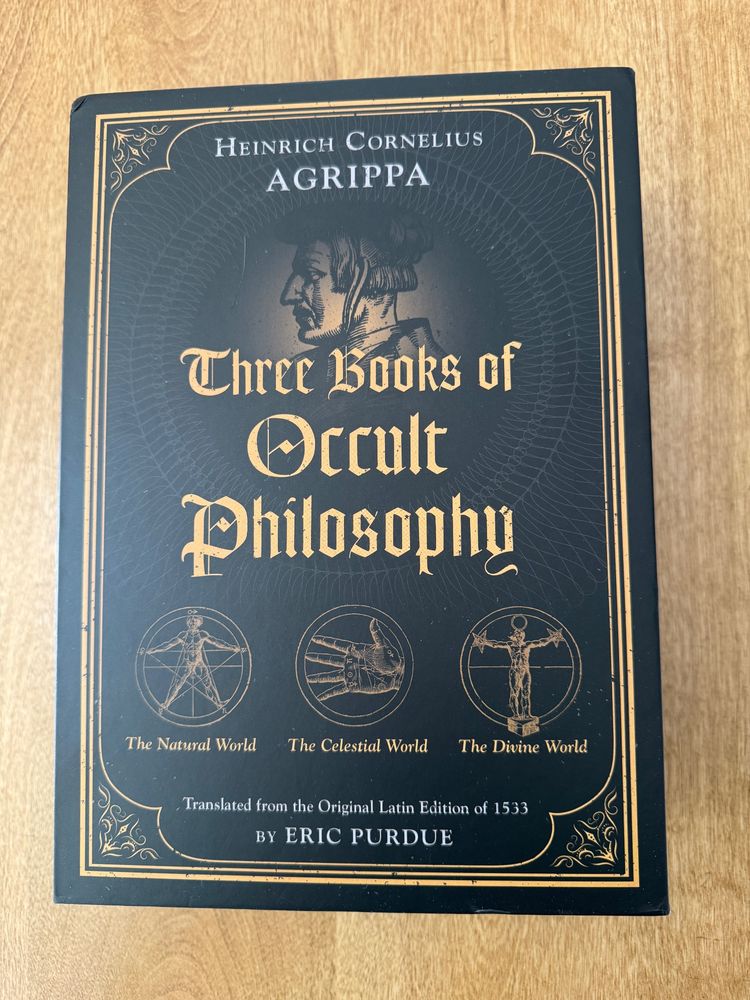 Photograph of the slipcase for a collection of books. It is black with a gold border and writing, and a profile of Agrippa in the background also with three round symbols for each of the three books in the collection titled Three Books of Occult Philosophy; The Natural World, The Celestial World, and The Divine World. There is a notation that this was translated from the Original Latin Edition of 1533 by Eric Purdue. 