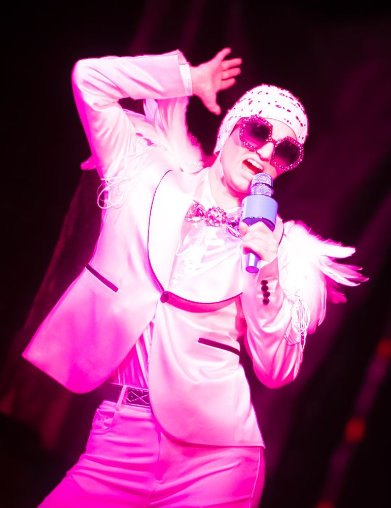 DragKing Apollo G. Accepted performs to an Elton John mix at a #Dragshow fundraiser at Vinos in #LittleRock.

Performer is standing on stage singing into a microphone under pink lights with a Feathered outfit and swim cap.