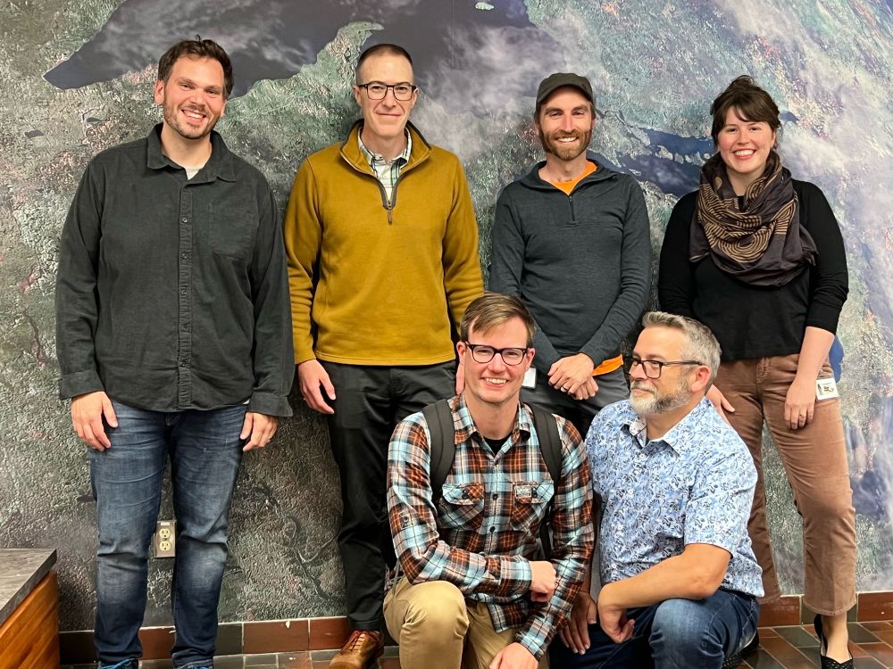 A group photo of five men and one woman in front of satellite image of great lakes. Everyone is smiling at camera.