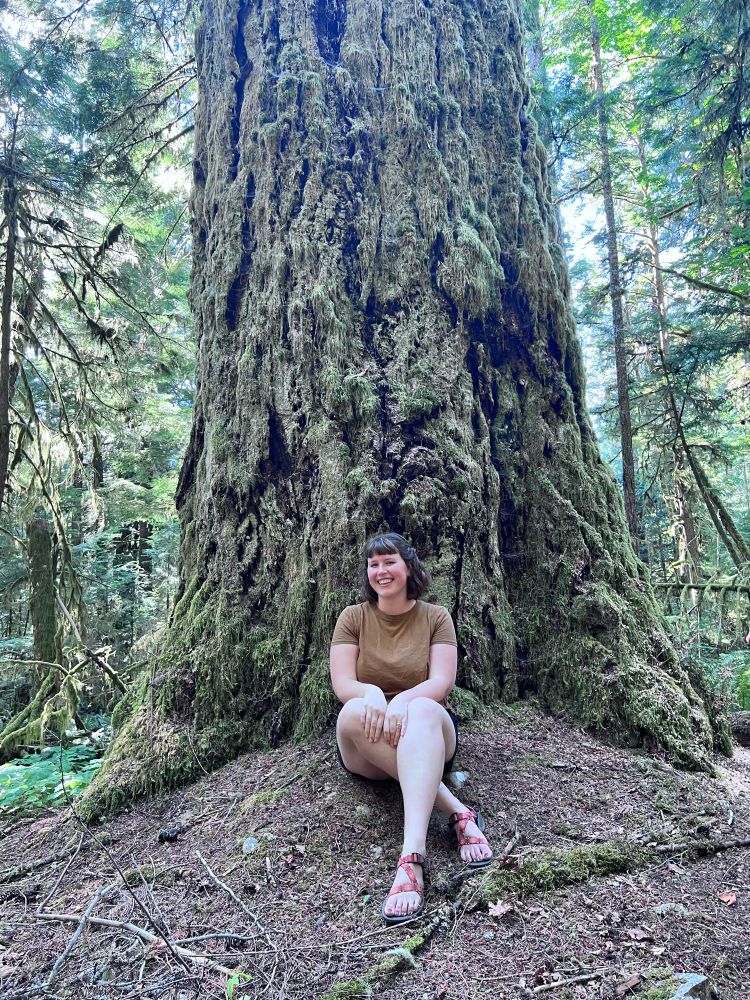 Smiling woman in brown shirt and sandals sits in front of large douglas fir tree covered in moss in a Vancouver Island forest.