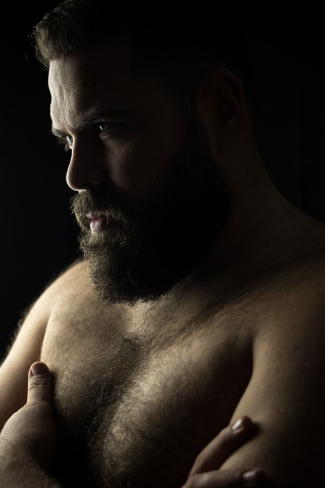 Retrato de un hombre con barba de perfil con los brazos cruzados. La luz lateral viene desde detrás del sujeto creando amplias sombras en buena parte de su rostro.