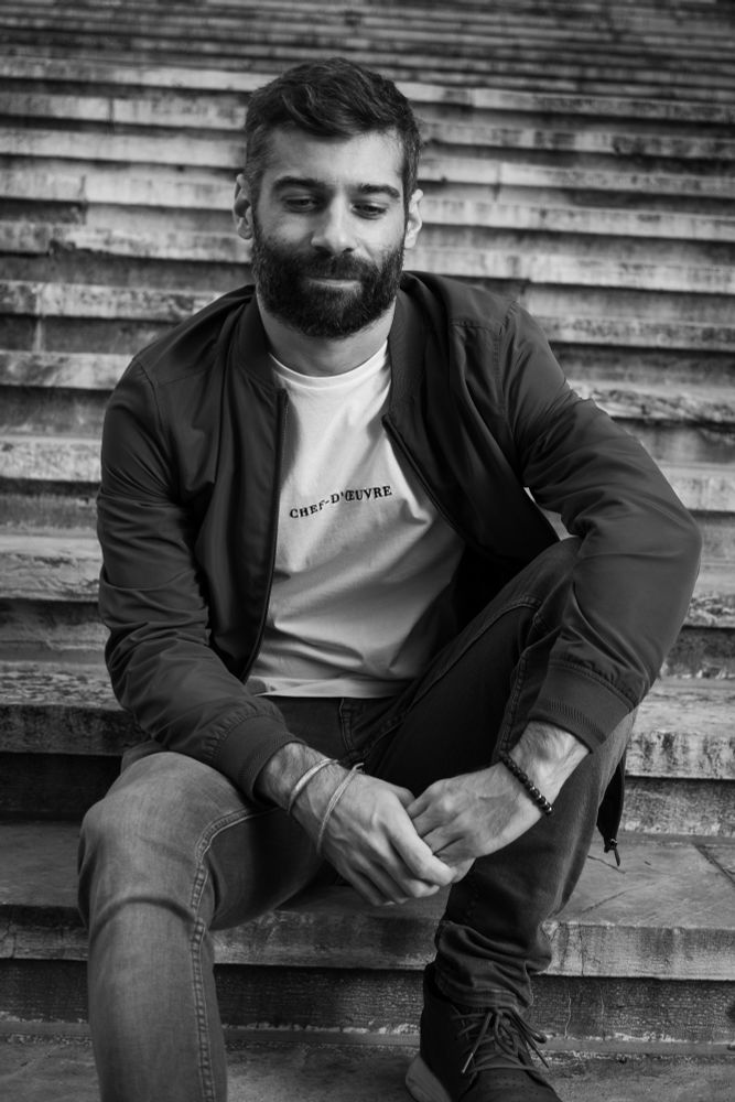 Foto en blanco y negro de un hombre joven con barba y chaqueta sentado en unas escaleras.