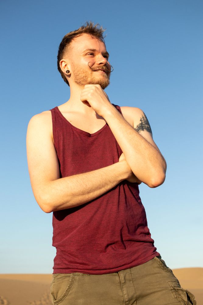 Hombre con bigote con camiseta de tirantes granate en las dunas con un cielo azul de fondo.