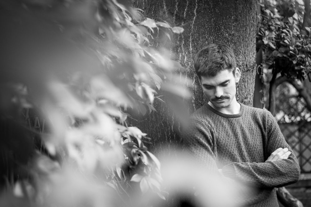 Foto en blanco y negro de hombre con jersey en apoyado en un árbol en la mitad derecha del encuadre, la mitad izquierda está dominada por un manto de hojas en distintos planos.