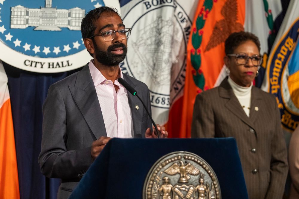 Council Member Shekar Krishnan speaking at a podium with flags behind at a Pre-Stated meeting. 