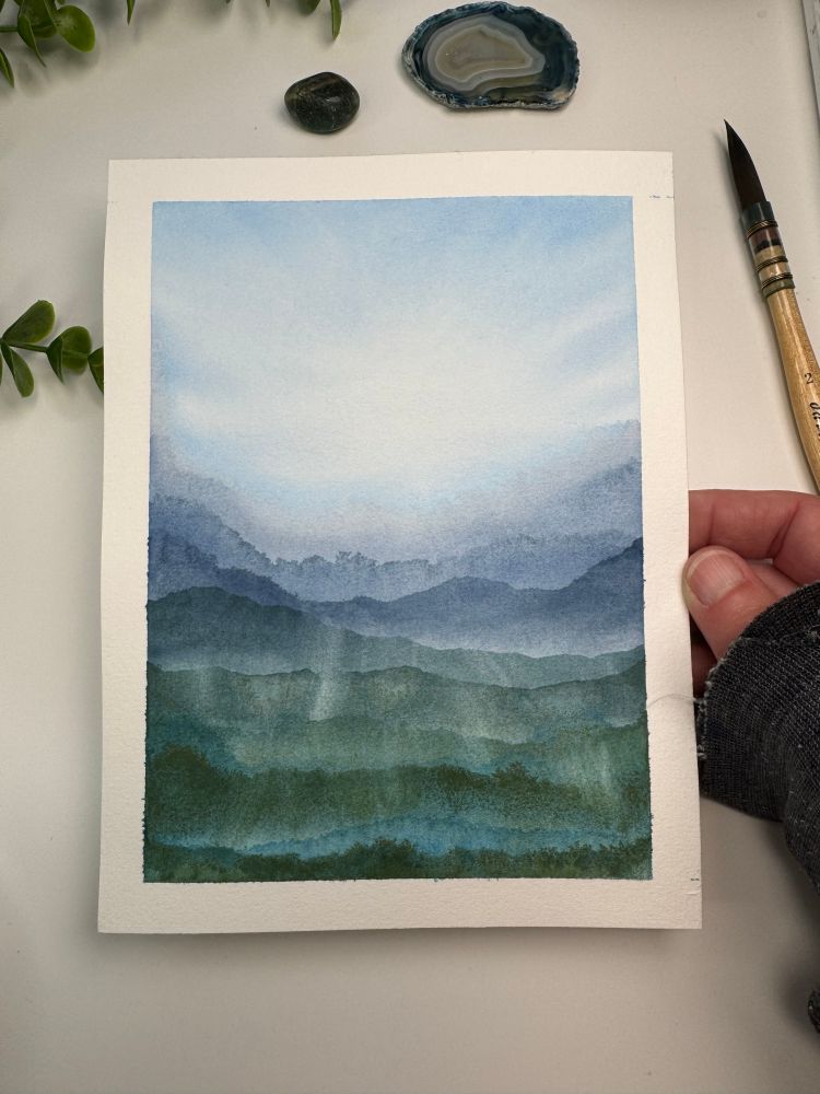 Watercolor painting of a blue and white sky with dark blue, green, and turquoise mountain layers.