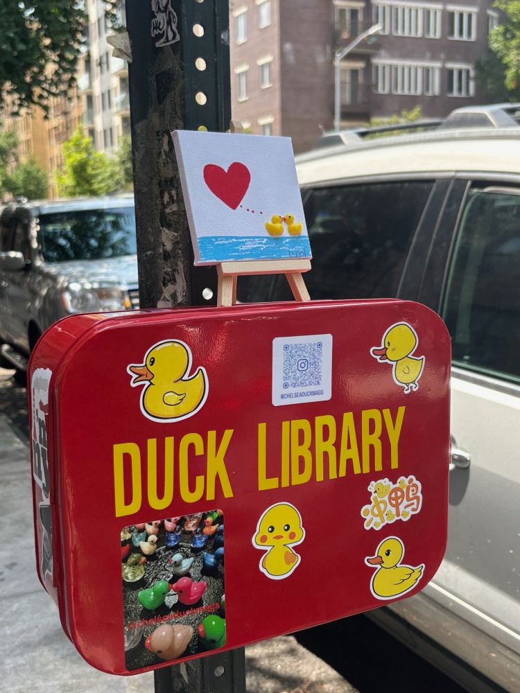 A red metal lunch box attached to a parking sign with the words DUCK LIBRARY in yellow, as well as various stickers, primarily of yellow rubber ducks. On top is a mini easel holding a canvas with a painting of two yellow rubber ducks on some water, with a red heart above them, presumably indicating their love for each other. While not pictured, the duck library contains various little ducks for people to collect and enjoy. 