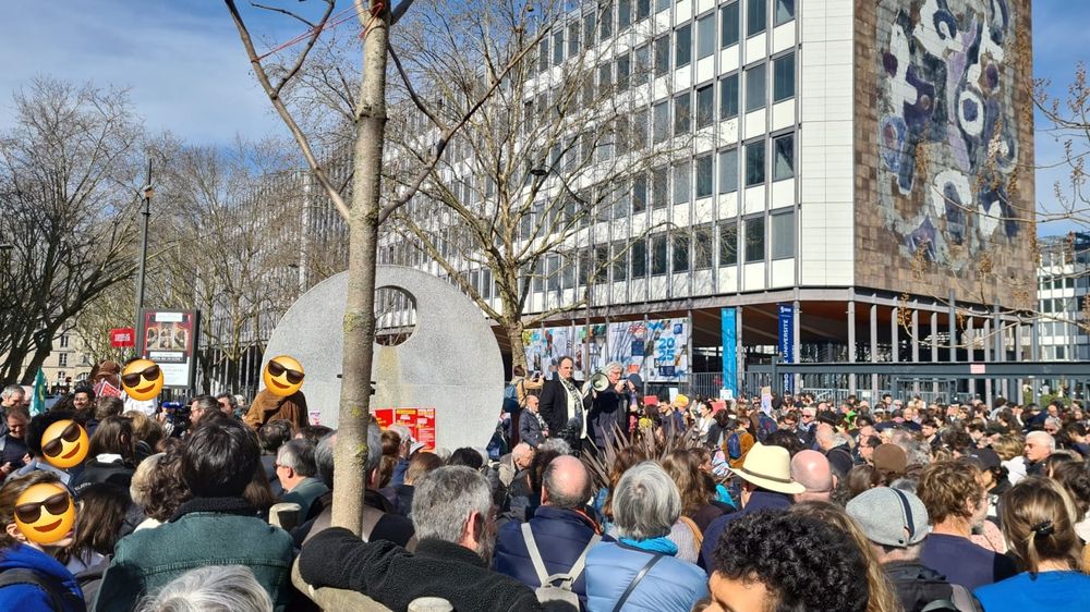 Vue de la place Jussieu couverte de monde en attendant le départ de la manifestation. On voit l'entrée du campus à droite, la fontaine au milieu, et certains visages sont couverts par des emojis sur la gauche. 