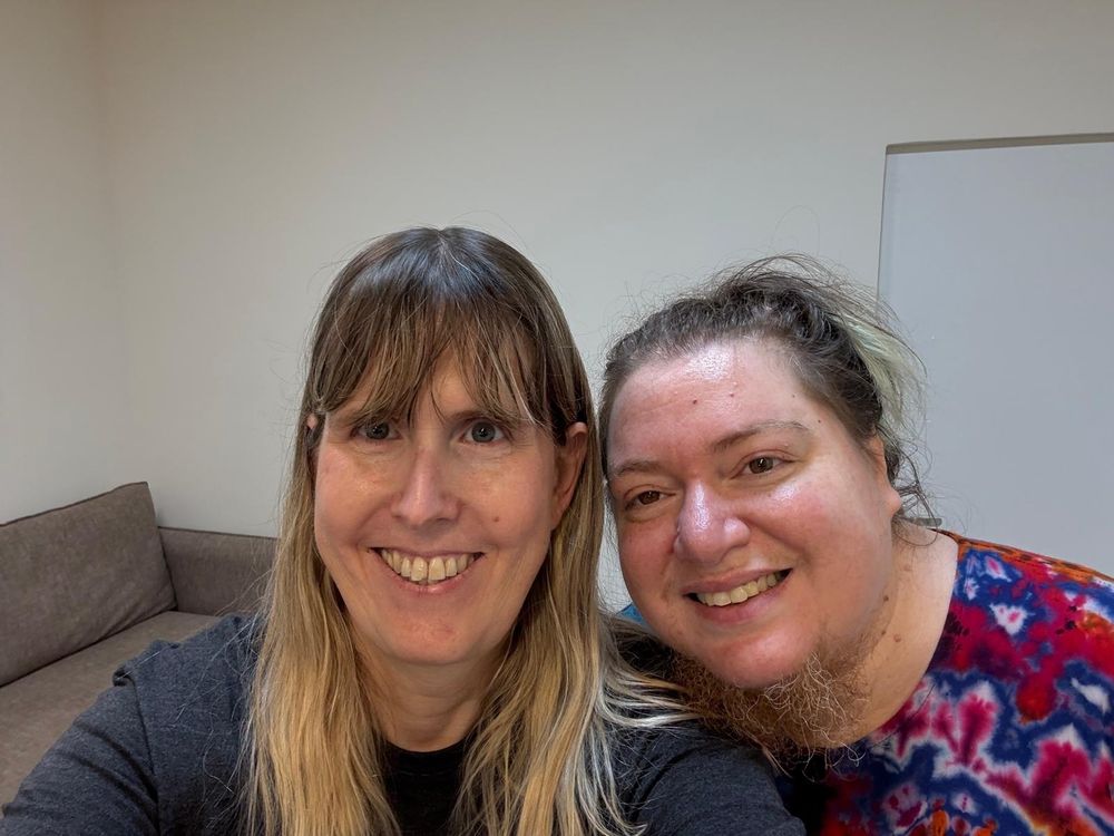 A thin blonde woman wearing a dark gray shirts and a fat brown hair person in a tie-dye dress smiling at the camera looking tired
