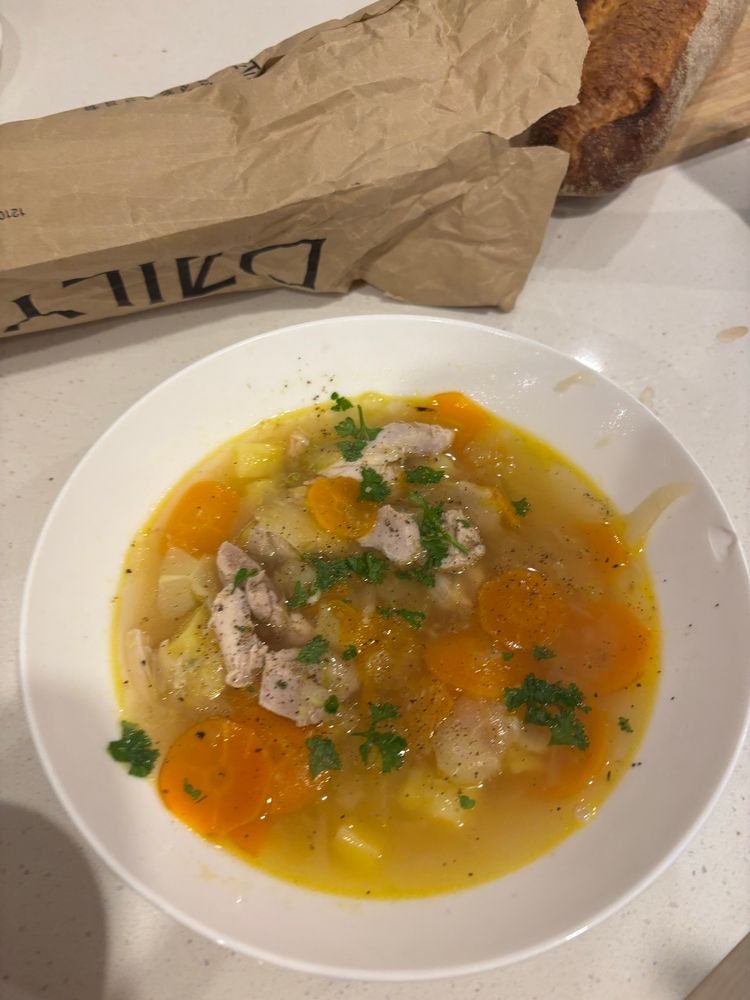 White bowl filled with chicken soup sitting in a counter with the edge of a baguette on a cutting board visible 