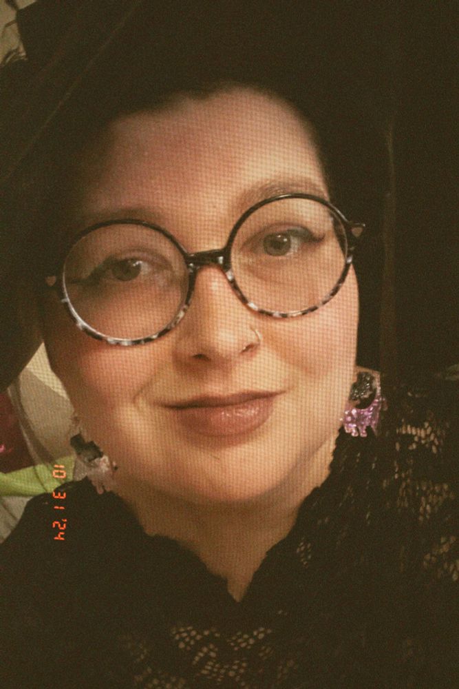 jude, a white woman with round black and tortoise shell glasses, is dressed for halloween in a large black hat with a veil, glittery purple moon earrings, and a lace dress.