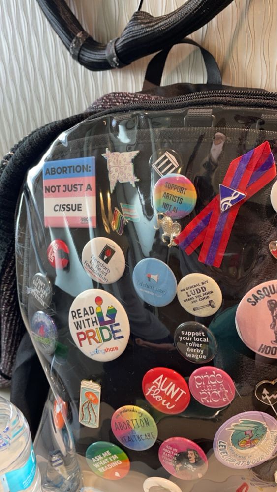 a black backpack covered in pins. some of the most prominent ones featured read “Abortion: Not Just a Cissue,” the internet archive logo, “Support Artists Not AI,” “No General But Ludd Means the Poor Any Good,” and “Read With Pride.”