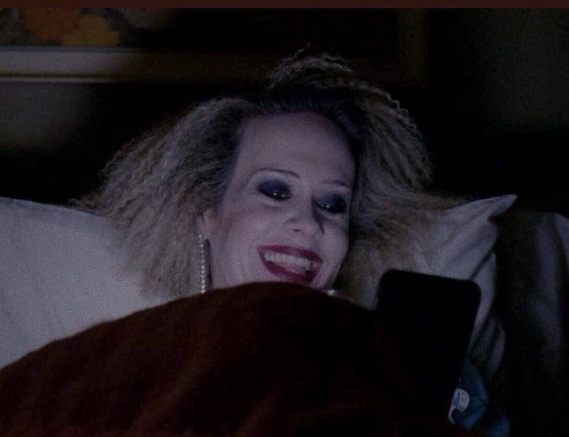 Hypodermic Sally from American Horror Story on her phone in bed, laughing.