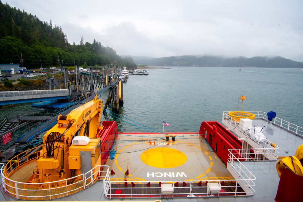 View from a ship’s deck toward a dock lined with boats on a misty, overcast day. The foreground shows a large yellow crane and a circular yellow winch area with the word 'WINCH' painted on it, while forested hills rise in the background.