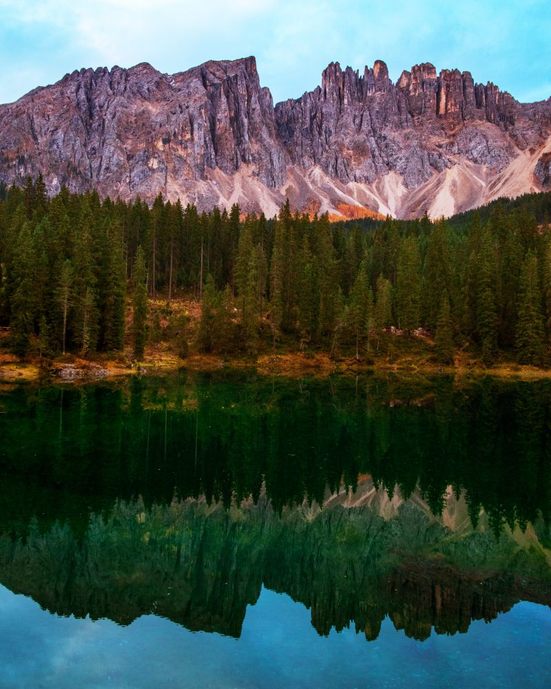 Jagged limestone cliffs rise sharply above a dense forest of evergreen trees, mirrored perfectly in the still, green waters of a mountain lake in the Dolomites. The composition captures the symmetry and serenity of the landscape at twilight.