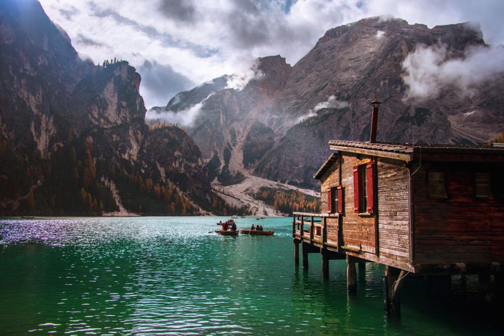 A rustic wooden boathouse on stilts juts out over the emerald waters of Lago di Braies in the Dolomites. In the distance, rowboats drift across the lake, surrounded by dramatic mountain cliffs and scattered autumn trees, under a sky of breaking clouds and light.