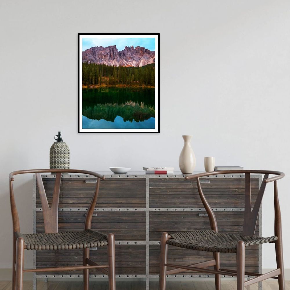 A framed photograph hangs above a rustic wood-and-metal sideboard, featuring a serene mountain scene from the Dolomites. The image shows jagged cliffs rising above a dense pine forest, reflected perfectly in the still green waters of a lake. The setting includes two modern wooden chairs and minimalist decor, blending natural and contemporary design elements.