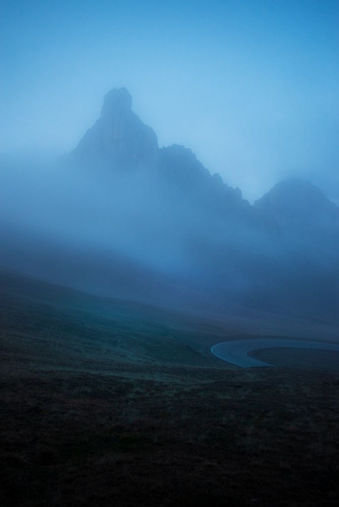 A winding mountain road curves through a mist-covered valley beneath a towering, craggy peak in the Dolomites. The scene is enveloped in deep blue fog, softening the rugged contours and creating a moody, atmospheric effect.