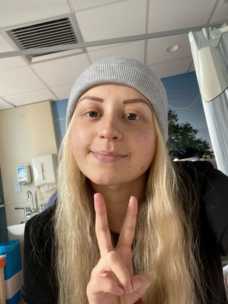 A young girl with long blonde hair poses for a selfie whilst in hospital receiving treatment for primary bone cancer. She is wearing a grey beanie and is showing the peace sign with her fingers.