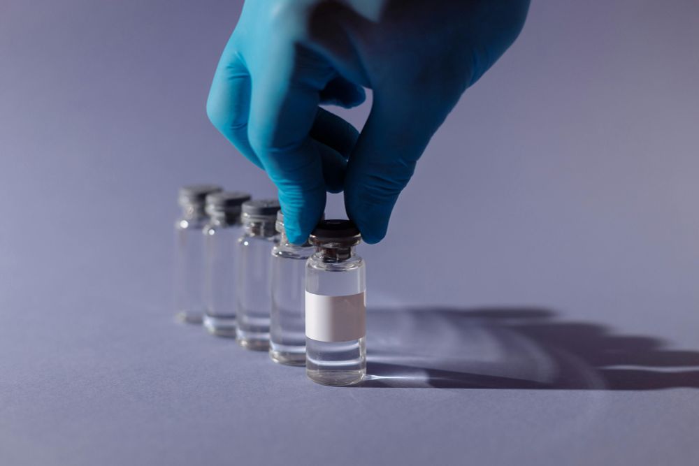 A hand, covered by a safety glove, reaches out to pick a specific bottle of medicine from a row of five.
