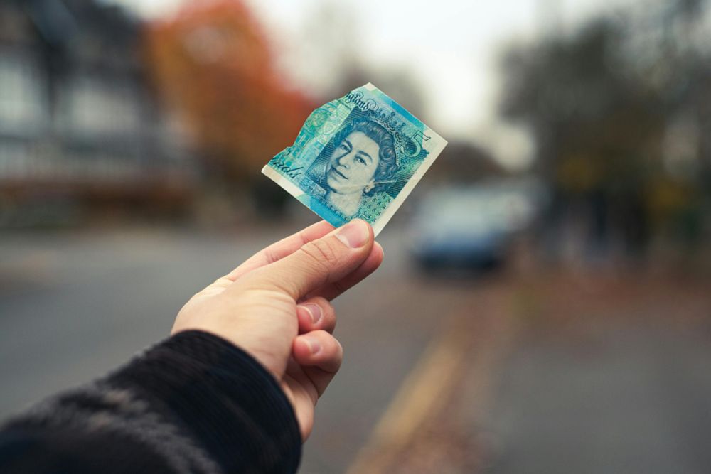 A photograph showing an outstretched handing holding a torn £5 note on a street with a blurred background. 