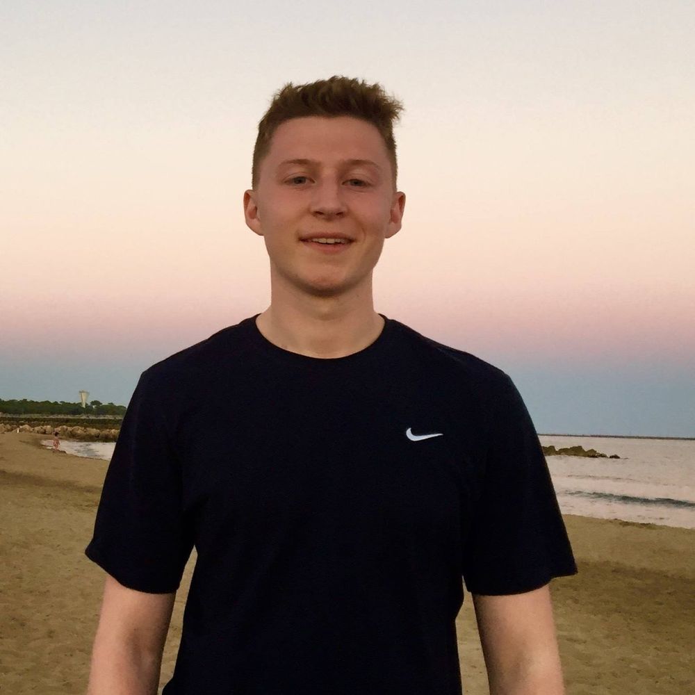 A young boy poses for a close-up photograph on a beach during sunset. He is wearing a black t-shirt and smiling at the camera.