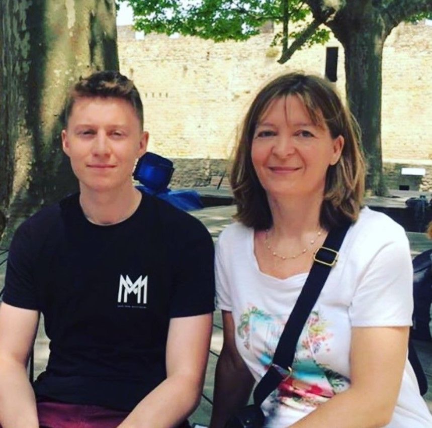A mother and son pose side-by-side for a photograph. They are both smiling at the camera and look relaxed whilst sitting on a bench.