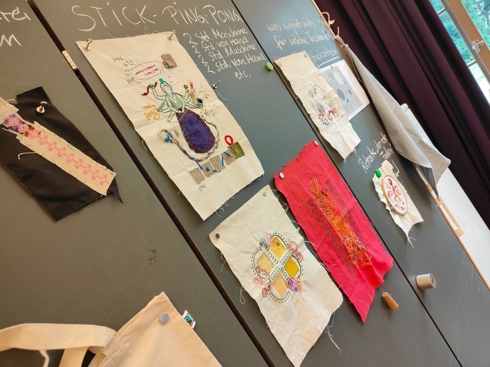 Photo of textile embroidery art pieces hanging up on wall with writing giving info about the pieces. One piece is a beige fabric with a many arm woman in a purple dress holding various things. Another is an embroidery design of swirls and squares. Another is embroidery on red fabric, of a yellow phoenix, surrounded by shibori like pattern embroidery
