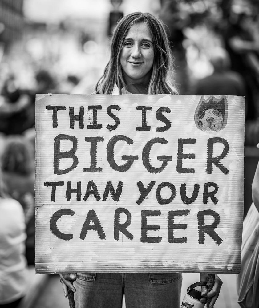 A black-and-white photo of a young woman at a London protest, smiling as she holds a large sign that reads in all caps, “This is bigger than your career.” Her hair falls just past her shoulders, and the sign covers her torso. The image captures a powerful, hopeful moment of protest and solidarity. Photo by Misan Harriman