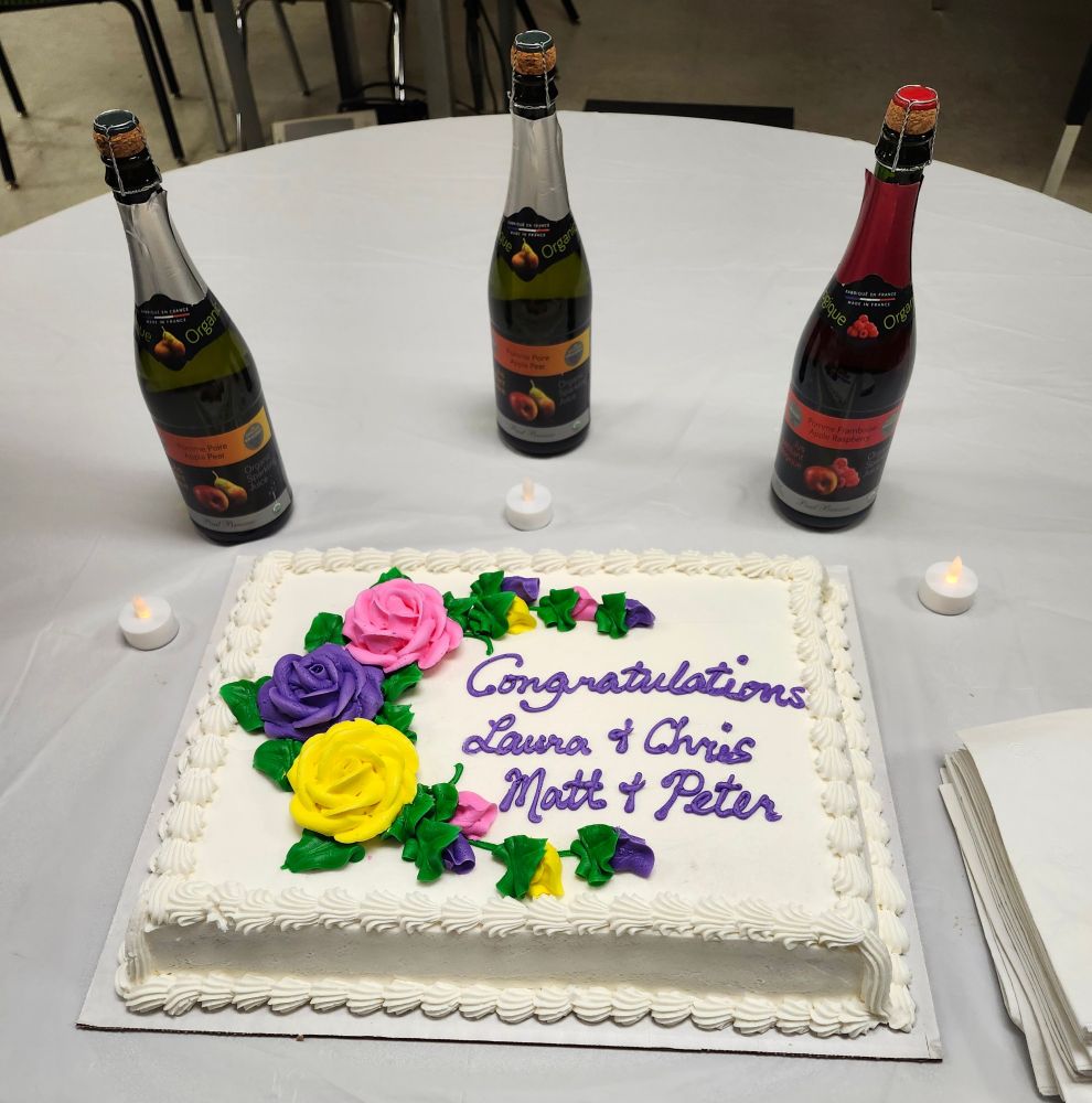 A white sheet cake with purple icing saying "Congratulations Laura & Chris, Matt & Peter" sits on a white table in front of three bottles of sparkling fruit juice.
