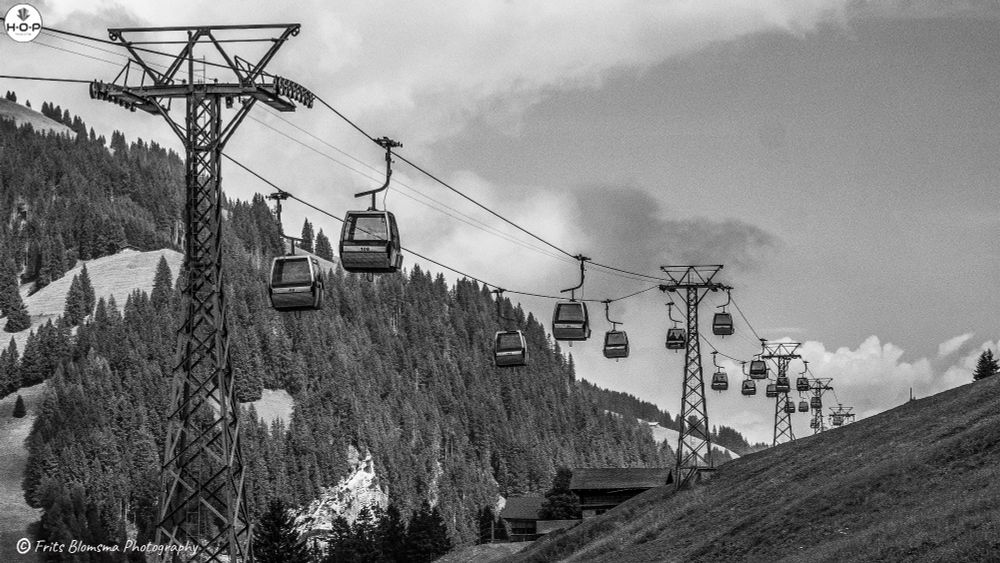This serene photograph captures a line of gondolas gliding gracefully over the lush alpine landscape a quintessential scene of the European mountains, likely in Switzerland or Austria. The cable cars ascend steadily toward the horizon, their rhythmic spacing drawing the eye along the line toward distant peaks. 

Below, rolling green pastures dotted with rustic wooden chalets give way to dense pine forests that cloak the slopes. The crisp blues of the sky and the deep greens of the hills form a soothing palette, while the metallic structure of the lift towers adds a contrasting note of industrial precision.

What makes this photograph compelling is its sense of quiet motion and harmony. The gondolas small capsules of human presence — seem almost to float in the stillness of the mountain air. The composition evokes both tranquility and anticipation: a peaceful moment before the next ascent, a gentle reminder of how technology can coexist with nature’s grandeur when designed thoughtfully.