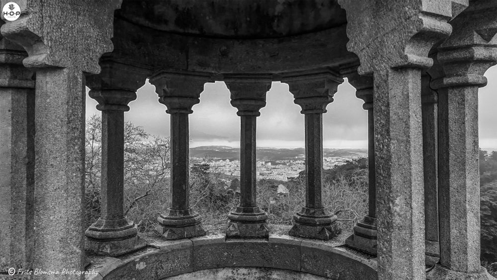 This photo captures a vibrant, eclectic palace built on a dramatic hilltop, blending several architectural styles into one striking panorama. The structure features ornate stonework, arched windows, tiled façades, and towers capped with golden domes. The contrast between the cool gray-and-white detailing on the left and the bold yellows and reds on the right creates a vivid, almost dreamlike composition. The mix of Gothic, Moorish, and Manueline-inspired elements gives the building a whimsical, fairy-tale quality. With the blue sky above and lush greenery below, the entire scene feels like a colorful fusion of history, art, and imagination rising out of the landscape.