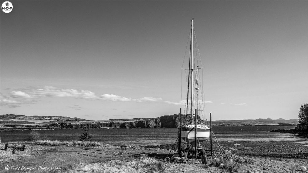 A still moment on the Isle of Skye: a lone sailboat rests on its cradle at the water’s edge, waiting for the next tide and the next adventure. The bay is calm under a cloudless blue sky, framed by rugged cliffs and gentle hills that stretch into the distance. Sunlight glints off the deep blue water, and the quiet shoreline feels untouched, suspended between sea and sky. It’s the kind of scene that captures the essence of Skye remote, peaceful, and quietly powerful, where even an idle boat seems to hold a story.