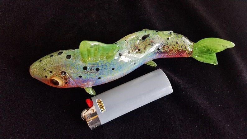 A rainbow trout bong/pipe with bright green, bright pink and transparent colors. There's a white lighter next to it and the bowl is in the mid back of the fish