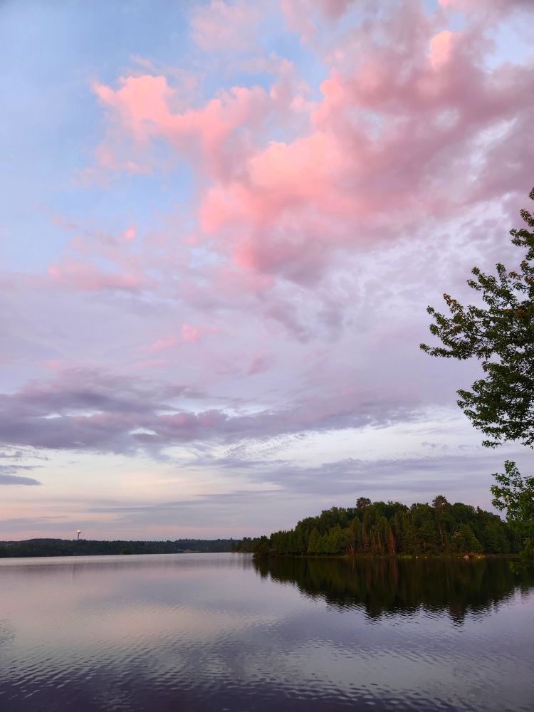 A lake scene with trees on the horizon and a light blue sky with fluffy pink clouds
