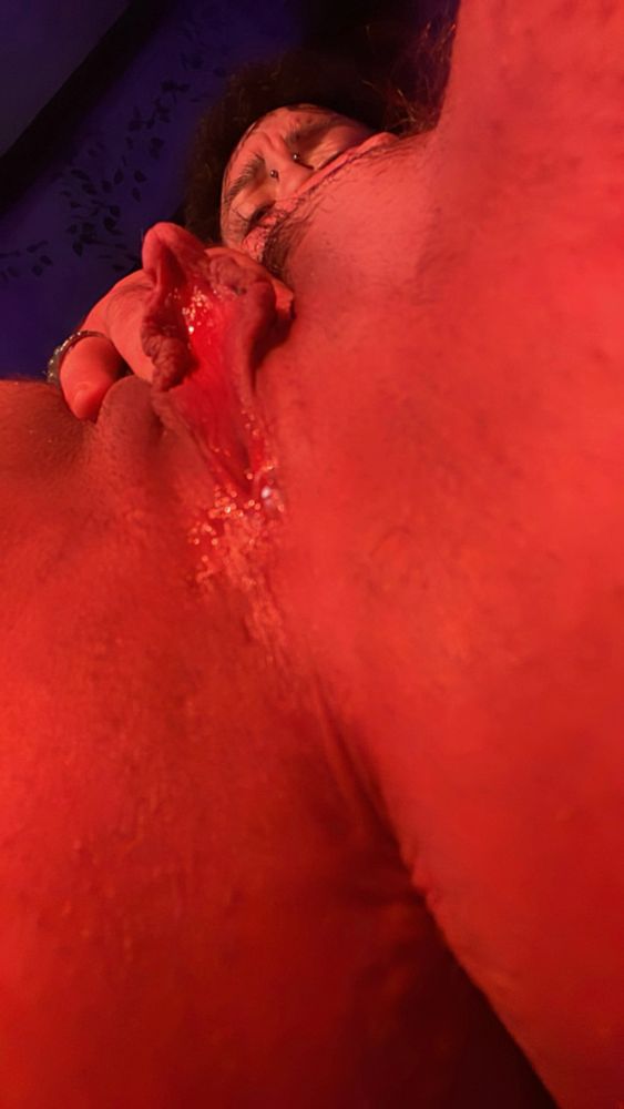 A low angle photo of J’s holes in warm orange lighting. His legs are open and you can just barely see his hand and furrowed brow at the top of the photo. He is pulling his clit back slightly to expose his glistening boy pussy.