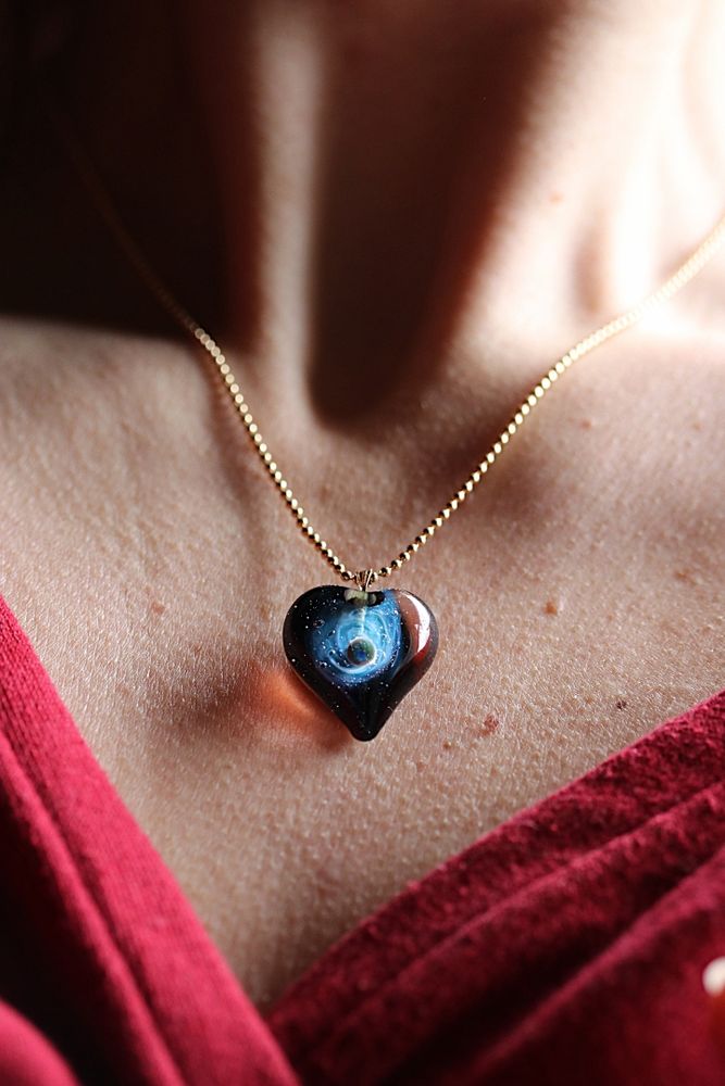 Collier en forme de cœur avec en son centre une galaxie de couleur bleu et une planète en apesanteur dans la voie lactée.

Le collier associée à une chaîne en or est porté sur le bust d'une femme.

A heart-shaped necklace with a blue galaxy and a weightless planet in the Milky Way at its center.

The necklace, paired with a gold chain, is worn across a woman's chest.