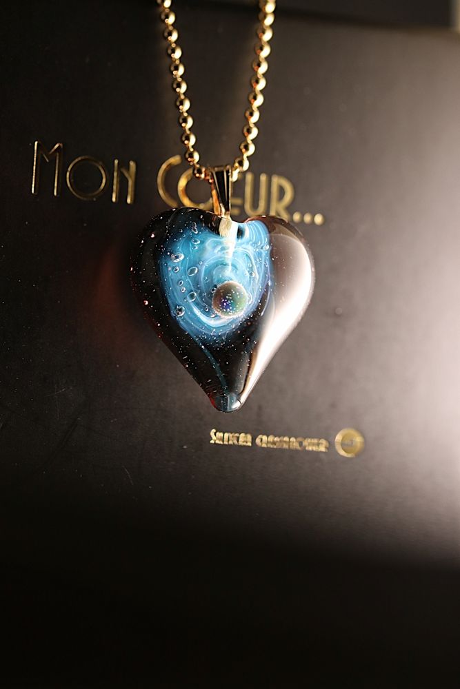 Collier en forme de cœur avec en son centre une galaxie de couleur bleu et une planète en apesanteur dans la voie lactée.
 Heart-shaped necklace with a blue galaxy and a weightless planet in the Milky Way in its center.