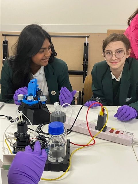 Two smiling students, one looking directly at the camera, with a Pioreactor and OpenFlexure microscope in front of them. There is an extension cord with a power supply next to it.
