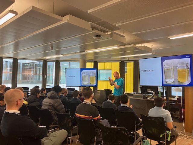Gerrit giving a presentation on biotechnology and sustainable protein to a group of students sitting on black chairs. There are two large screens showing the presentation.