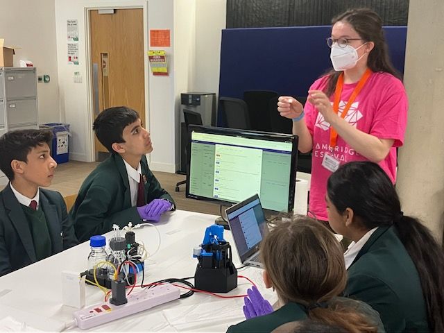 Sandrine, an AMYBO volunteer, explaining how the Pioreactor works to a group of students sitting around a white table. There is a Pioreactor, an OpenFlexure microscope, a laptop and a screen on the table.