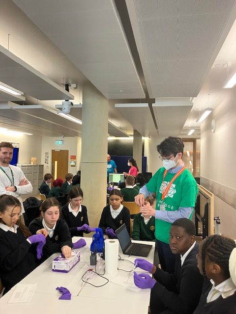 Rob, an AMYBO volutneer, handling a vial from the Pioreactor, surounded by a group of students sitting at a white table. There is a Pioreactor, laptop, kitchen roll and a box with purple disposable gloves on the table.