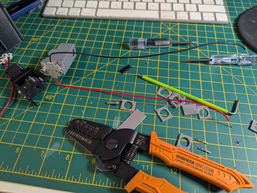 A bunch of 3D printed components, wires and screws are lying on a cutting mat. There's an orange Engineer PA-09 crimping tool in the foreground, as well as some screwdrivers and an allen key.
