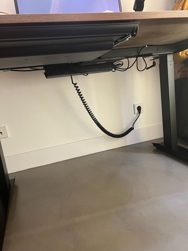 A photo of a desk underneath, with a white wall and a curly/spring-like cable coming from the desk surface into a socket on the wall 