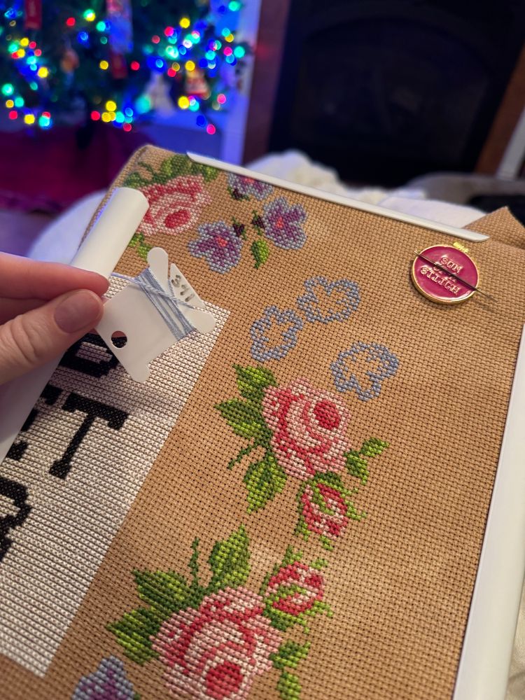 My almost finished cross stitch project of a sign from the Disney World ride the Haunted Mansion. It is just a corner with flowers. At the top are blue flowers with no shading or leaves stitched in yet. To the left I am holding a DMC thread bobbin with very short strands of the blue.