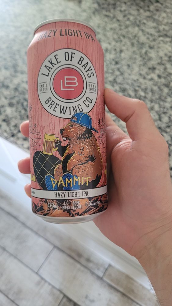 A pink can from Lake of Bay's brewing. It has a logo of a Beaver wearing a blue ball cap backwards, drinking a beer. The beer name is called Dammit, it is a hazy light IPA.