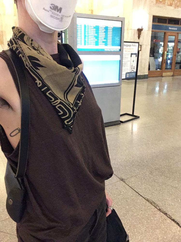 Torso shot of a skinny white person wearing a dark-brown baggy tank top and a beige-and-black bandana. Only the bottom half of the face is in frame, wearing a 3M Aura N95 mask.