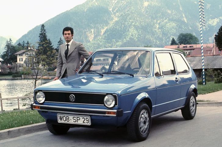 A young Giorgetto Giugiaro standing next to a blue Volkswagen Golf mk1, car he designed.