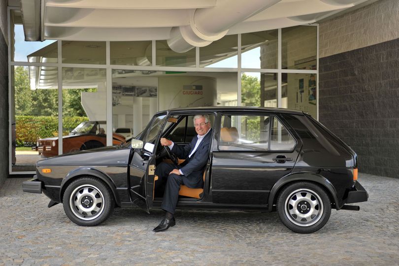 Giorgetto Giugiaro sitting in the driver's seat of a Volkswagen Golf mk1, a car he designed, with the door open and one leg on the floor.