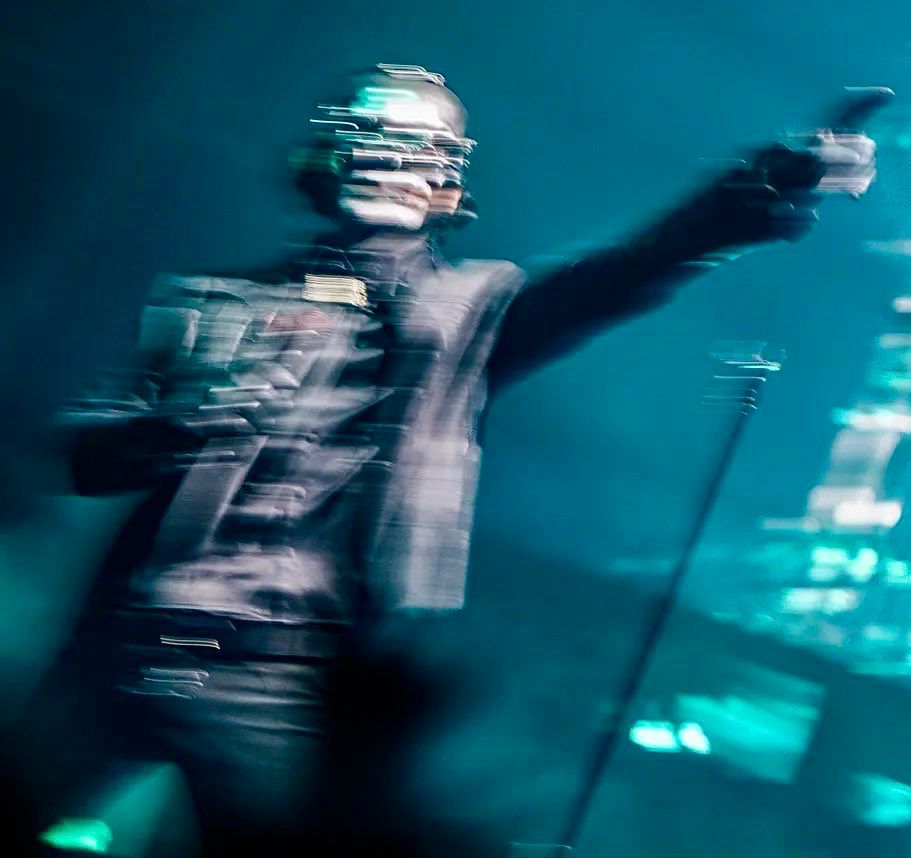 Blurred image of Papa V Perpetua in his leather jacket ensemble with his left hand pointing sideways. The backdrop is in blue-green lighting